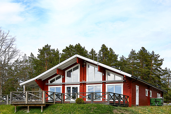 En röd reception- och restaurangbyggnad med stora fönster och altan, omgiven av grön natur och tallskog.